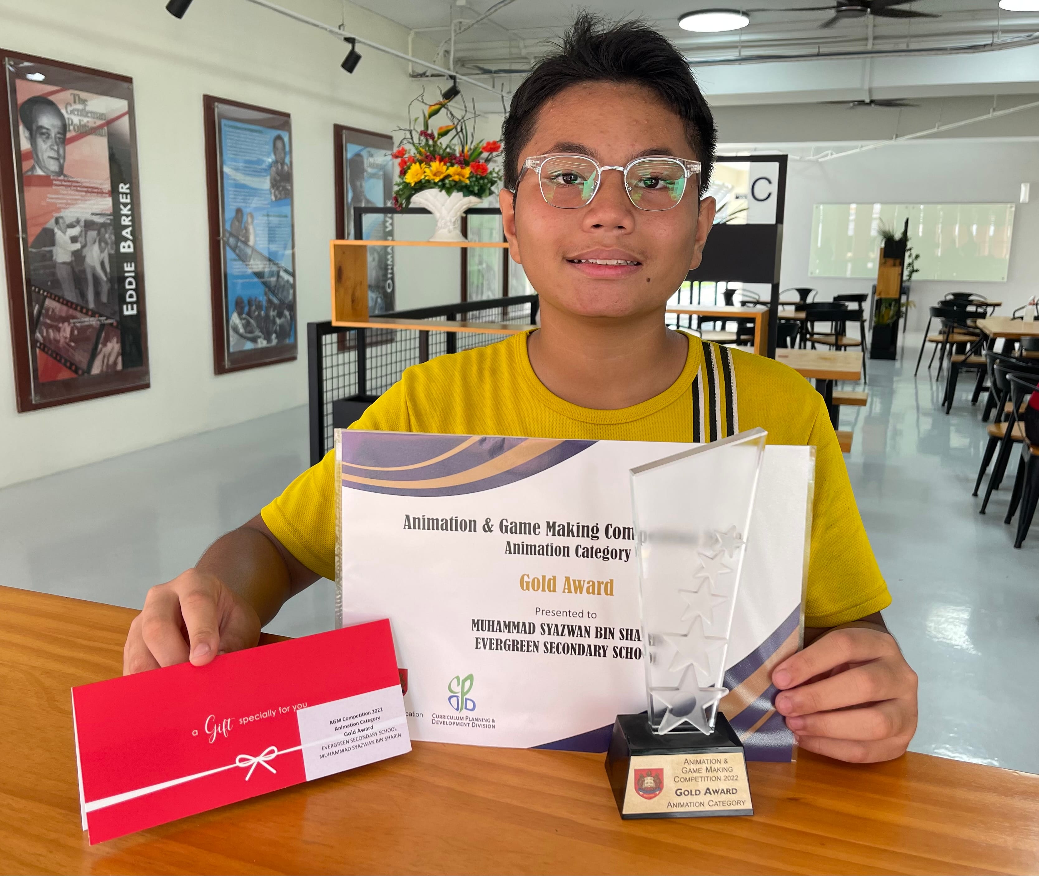 A young boy wearing glasses holds a Gold Award certificate, trophy, and gift envelope from an Animation and Game Making Competition.